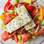 A bowl of Authentic Greek Salad with sliced tomatoes, cucumbers, red onions, red bell peppers, olives, and two slabs of feta cheese, all topped with olive oil and herbs.