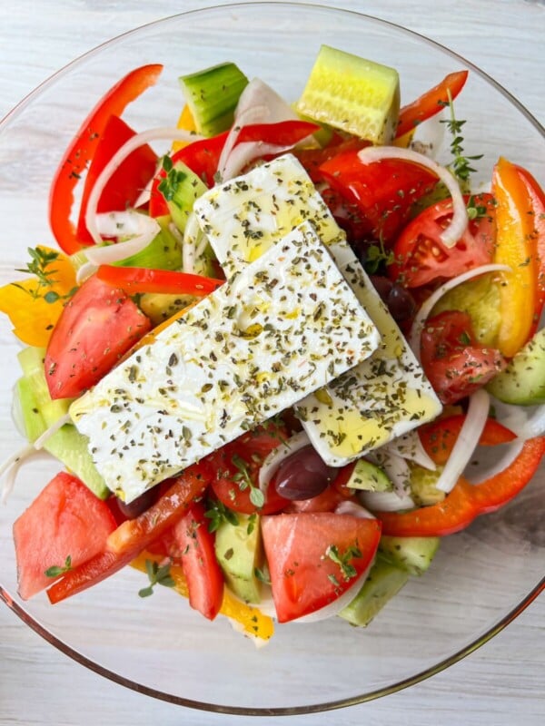 A bowl of Authentic Greek Salad with sliced tomatoes, cucumbers, red onions, red bell peppers, olives, and two slabs of feta cheese, all topped with olive oil and herbs.