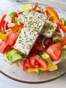 A bowl of Greek salad, similar to Maroulosalata, features sliced tomatoes, cucumbers, red and yellow bell peppers, onions, olives, and two large pieces of feta cheese topped with herbs.