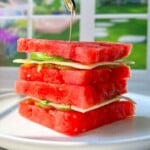 A refreshing Watermelon Sandwich, featuring layers of watermelon, cheese, and greens held together by a toothpick, sits on a white plate with a blurred garden visible outside the window in the background.