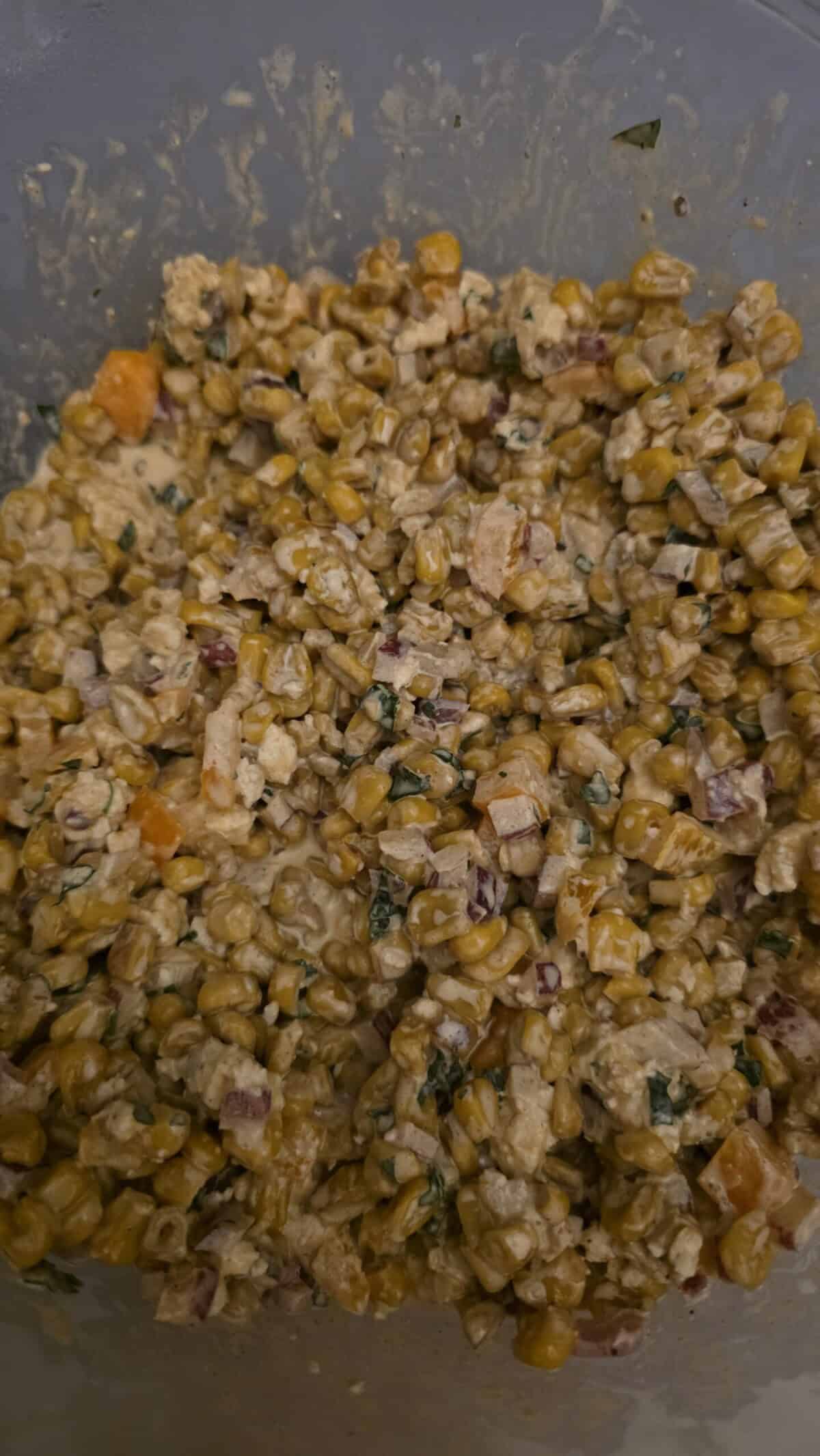 A bowl filled with a creamy corn salad mixture, including corn kernels, chopped red onion, herbs, and small pieces of orange vegetable, all coated in a sauce or dressing.