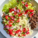 A Beef Burrito Bowl Recipe comes to life with shredded lettuce, diced tomatoes, cucumbers, red onions, cooked ground beef, and spaghetti noodles, all artfully arranged in sections on a white surface with sunlight streaming in.