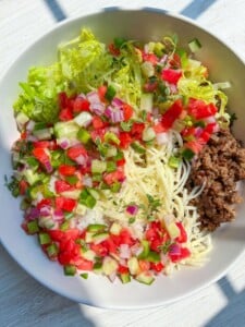 A Beef Burrito Bowl Recipe comes to life with shredded lettuce, diced tomatoes, cucumbers, red onions, cooked ground beef, and spaghetti noodles, all artfully arranged in sections on a white surface with sunlight streaming in.