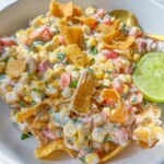 A bowl of creamy Frito Corn Salad with diced tomatoes, chopped herbs, crunchy corn chips, and a halved lime, served with a gold spoon on a white plate.