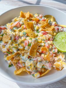 A bowl of creamy Frito Corn Salad with diced tomatoes, chopped herbs, crunchy corn chips, and a halved lime, served with a gold spoon on a white plate.