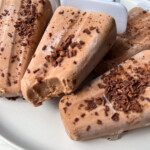 Close-up of chocolate popsicles made with a creamy Fudgesicle recipe, topped with chocolate shavings and arranged on a white plate. One popsicle has a bite taken out, revealing its rich, smooth texture inside.