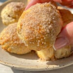 A hand holds a round, golden-brown Banana Donut Hole coated with sugar, above a ceramic plate filled with similar pastries on a light wooden surface—perfect for enjoying easy 2 Ingredient banana donut holes.
