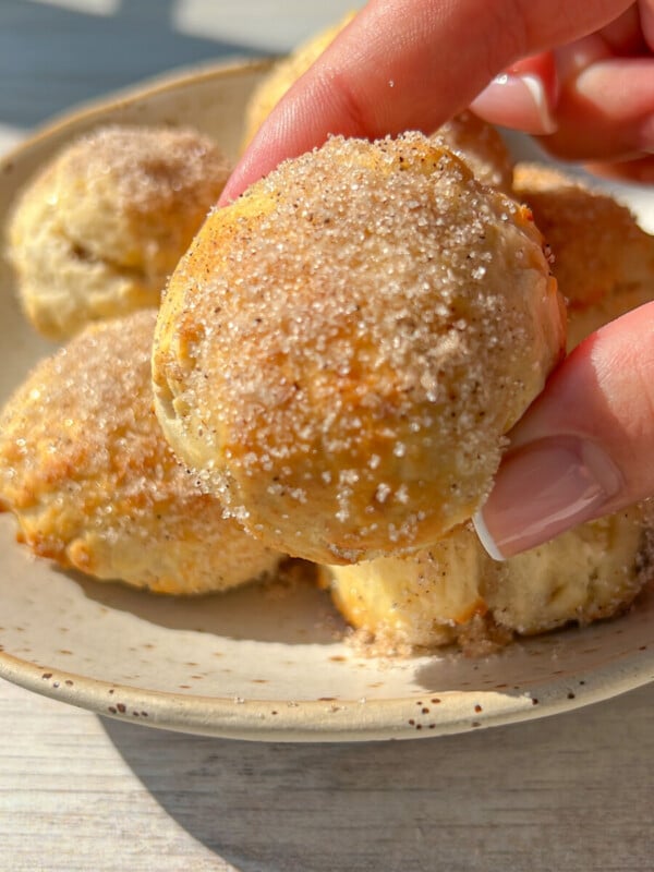 A hand holds a round, golden-brown Banana Donut Hole coated with sugar, above a ceramic plate filled with similar pastries on a light wooden surface—perfect for enjoying easy 2 Ingredient banana donut holes.