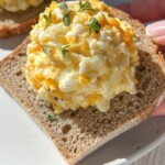A hand holds a slice of bread topped with creamy egg salad and fresh herbs, over a white plate on a light table—perfect for pairing with 4 Ingredient Pumpkin Donut Holes for a delightful meal or snack.