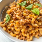 A close-up of macaroni pasta mixed with ground meat in a tomato-based sauce, reminiscent of classic Hamburger Helper, garnished with fresh basil leaves and served in a white bowl with a spoon.