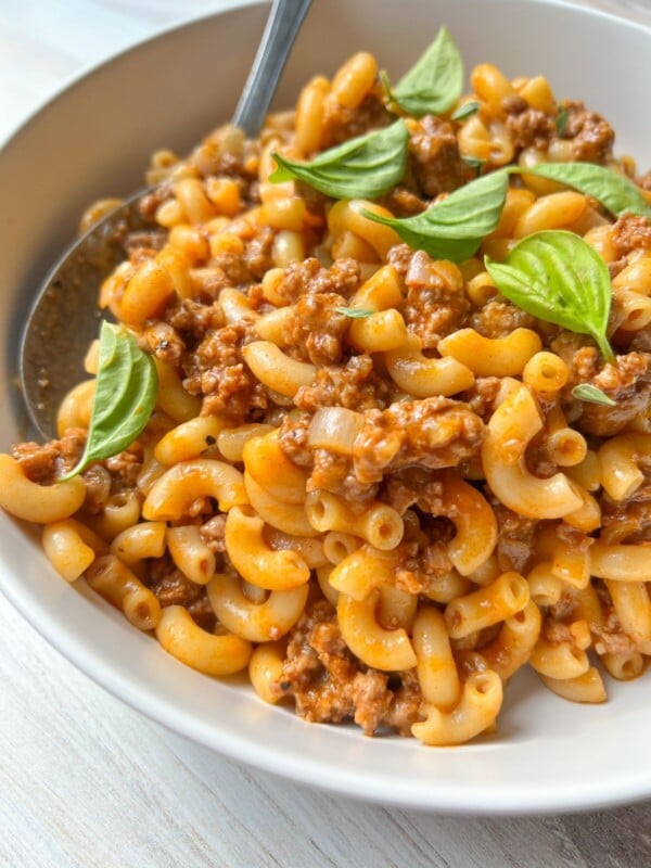 A close-up of macaroni pasta mixed with ground meat in a tomato-based sauce, reminiscent of classic Hamburger Helper, garnished with fresh basil leaves and served in a white bowl with a spoon.