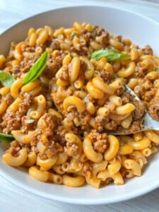 A bowl of macaroni pasta with ground beef in a creamy tomato sauce, reminiscent of homemade Hamburger Helper, garnished with fresh basil leaves. A spoon rests in the bowl, ready to eat.