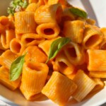 A close-up of rigatoni pasta covered in a creamy marinara sauce recipe, garnished with fresh basil leaves, served in a white bowl on a light wooden surface.