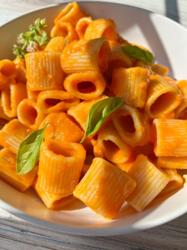 A close-up of rigatoni pasta covered in a creamy marinara sauce recipe, garnished with fresh basil leaves, served in a white bowl on a light wooden surface.