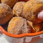 A hand holding a round, cinnamon sugar-coated 4 Ingredient Pumpkin Donut Hole hovers above a bowl filled with similar pastries. Sunlight highlights the golden brown donut holes inside the orange-rimmed bowl.