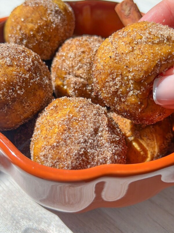 A hand holding a round, cinnamon sugar-coated 4 Ingredient Pumpkin Donut Hole hovers above a bowl filled with similar pastries. Sunlight highlights the golden brown donut holes inside the orange-rimmed bowl.