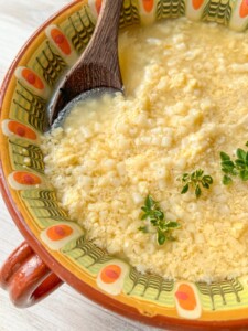A bowl of Stracciatella Soup with small pasta, garnished with fresh herbs. The bowl features a colorful patterned rim, and a wooden spoon rests inside the comforting soup.