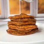A stack of golden brown 2 Ingredient Pancakes on a white plate, with syrup being poured from a metal pitcher, set in front of a window with a blurred autumn background.