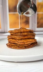 A stack of golden brown 2 Ingredient Pancakes on a white plate, with syrup being poured from a metal pitcher, set in front of a window with a blurred autumn background.