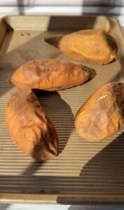Four whole sweet potatoes with rough orange-brown skin are placed on a ridged baking sheet, lit by natural sunlight—perfect for transforming into 2 Ingredient Pancakes or pairing alongside your favorite pancakes.