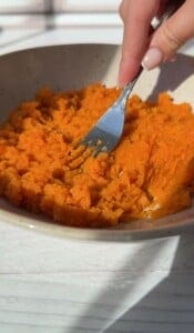 A hand holding a fork mashes cooked sweet potatoes in a shallow beige bowl on a white surface, with sunlight casting shadows—perfect prep for making easy 2 Ingredient Pancakes.