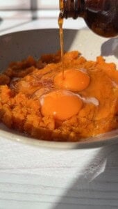 A close-up of two raw eggs cracked over mashed orange sweet potatoes in a bowl, as a brown bottle pours vanilla extract onto the mixture—perfect for making simple 2 Ingredient Pancakes. Sunlight highlights the ingredients.
