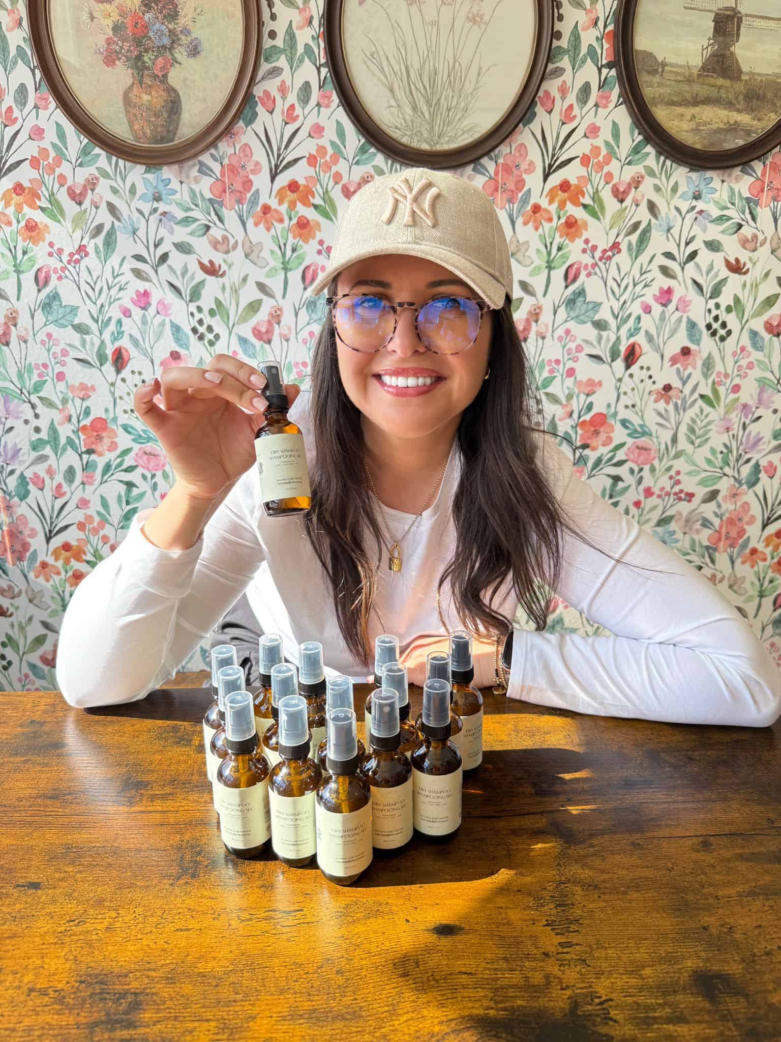 A person wearing a cap and glasses sits at a table holding a small bottle. In front are several identical bottles arranged in rows. The background features floral-patterned wallpaper and framed pictures. The person is smiling.