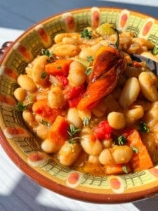 A colorful ceramic bowl filled with white beans, diced tomatoes, carrots, herbs, and topped with a roasted pepper, this vibrant bean stew is beautifully highlighted by sunlight.