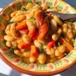 A colorful bowl of Bean Stew with white beans, carrots, red peppers, and herbs, topped with a roasted chili pepper and served in a decorative bowl on a sunlit table.