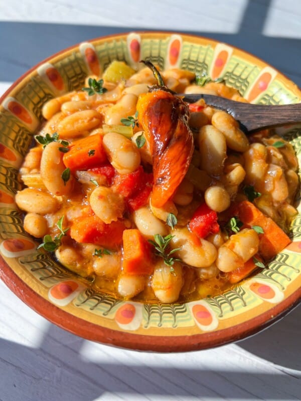 A colorful bowl of Bean Stew with white beans, carrots, red peppers, and herbs, topped with a roasted chili pepper and served in a decorative bowl on a sunlit table.