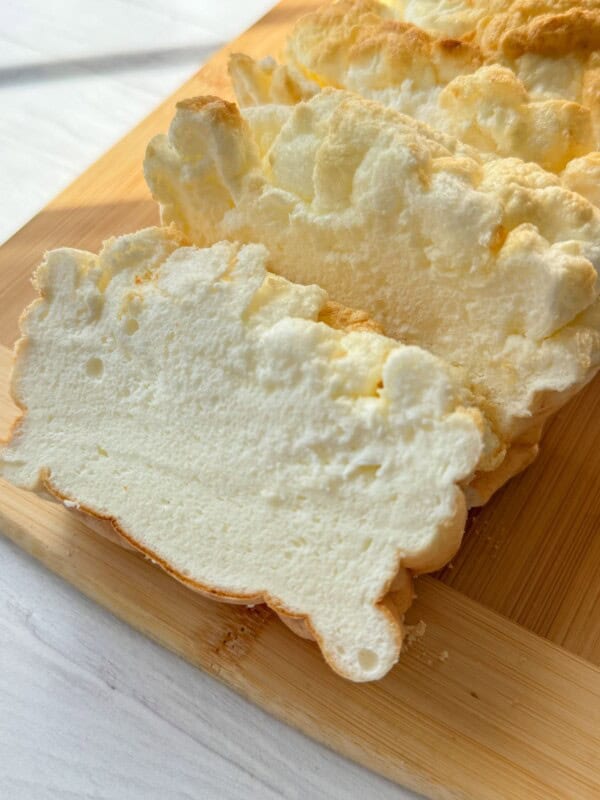 Sliced white, fluffy bread with a slightly golden crust sits on a wooden cutting board, showcasing its airy, cloud-like texture—perfect for trying your favorite Cloud Bread Recipe.