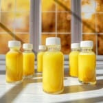 Six small glass bottles filled with bright yellow liquid, each sealed with a white cap, are arranged on a sunlit white surface. They’re perfect for trying your favorite ginger shots recipe against a warm yellow and orange autumn backdrop.