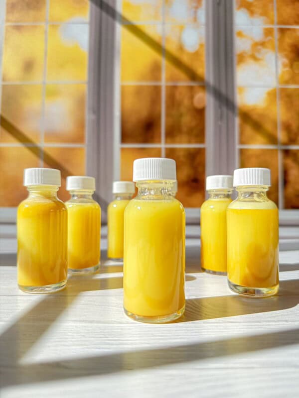 Six small glass bottles filled with bright yellow liquid, each sealed with a white cap, are arranged on a sunlit white surface. They’re perfect for trying your favorite ginger shots recipe against a warm yellow and orange autumn backdrop.
