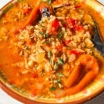 A bowl of hearty Stuffed Pepper Soup with ground meat, rice, chopped tomatoes, onions, herbs, and small peppers, garnished with fresh thyme. The soup features a rich red-orange color and a satisfying texture.