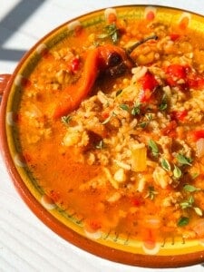A close-up of a bowl of Stuffed Pepper Soup with rice, ground meat, tomatoes, red peppers, a whole chili, and fresh herbs in a colorful ceramic bowl on a white surface.