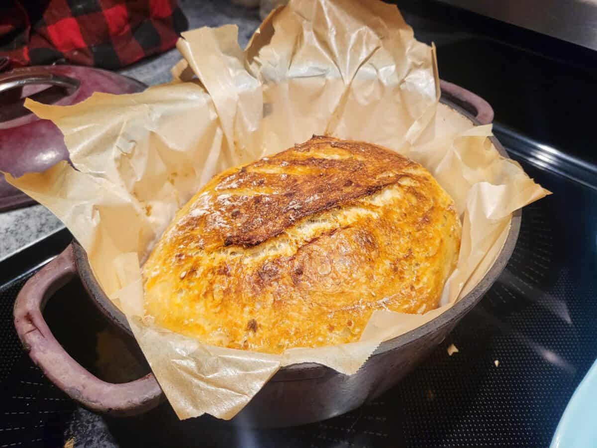 A round loaf of No Knead Feta Bread, crusty and golden-brown, sits in a parchment-lined cast iron pot on the stovetop, fresh from baking.