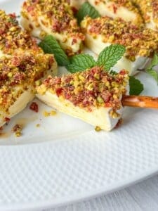 Slices of creamy cheese topped with crushed pistachios and dried fruit are arranged on a white plate. Fresh mint leaves are scattered for garnish, and a pretzel stick is used to pick up one of the slices.