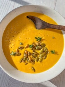 A bowl of creamy orange soup garnished with pumpkin seeds and small green herbs, served with a wooden spoon on a light wooden table.