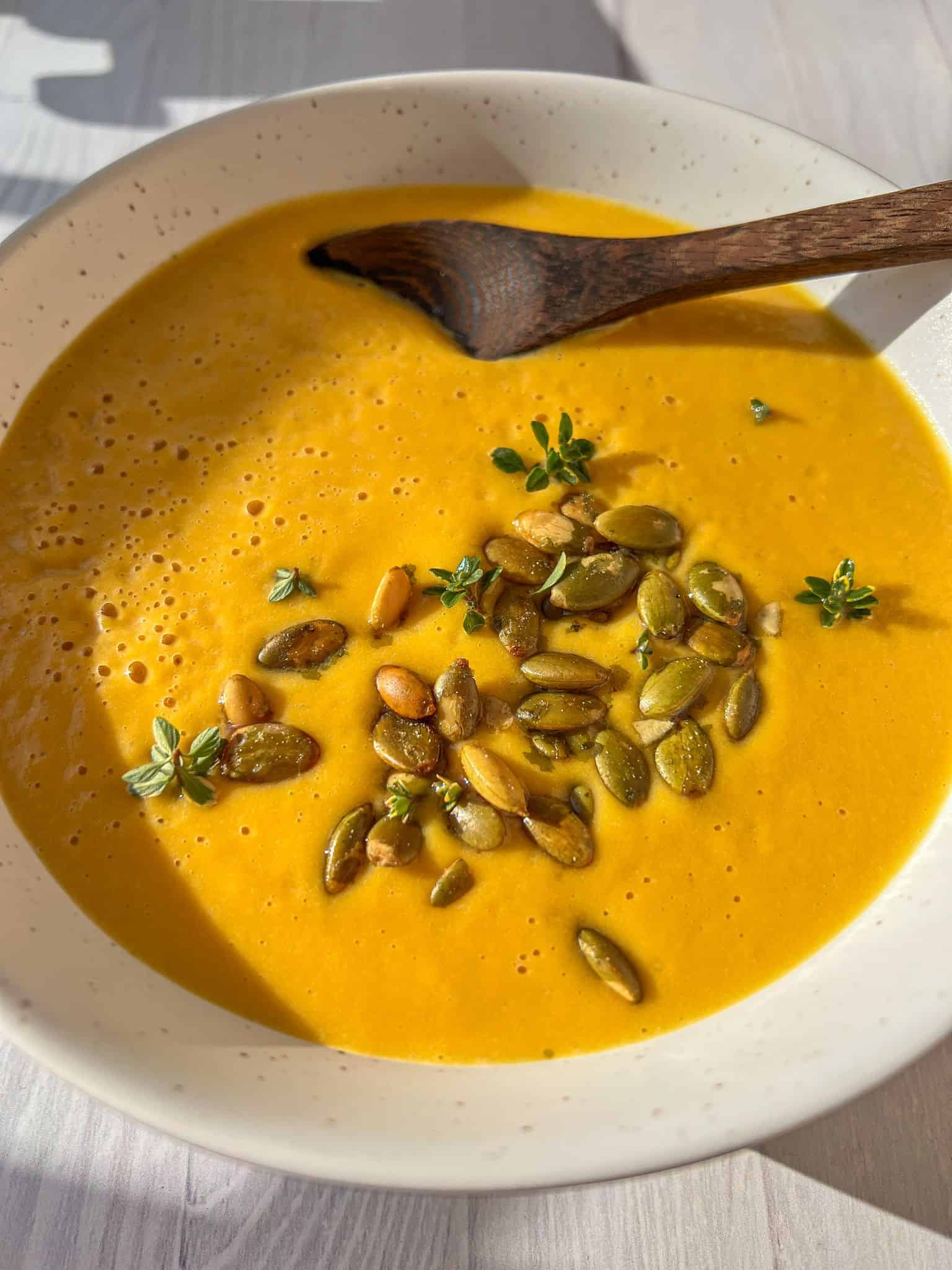 A creamy bowl of butternut squash soup garnished with pumpkin seeds and fresh thyme, served in a white bowl with a wooden spoon. Sunlight casts a natural shadow on the light-colored surface. - 6