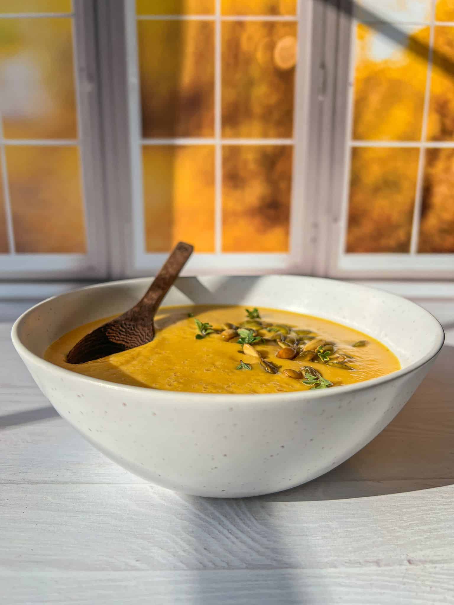 A bowl of creamy pumpkin soup garnished with herbs and seeds, placed on a white surface. A wooden spoon rests inside the bowl. The background features a window with a view of autumnal trees and vibrant sunlight streaming in. - 3
