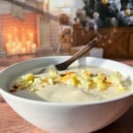 A bowl of creamy soup topped with roasted cauliflower sits on a wooden table. In the background, a cozy room features a decorated Christmas tree and a lit fireplace adorned with candles and seasonal decor.