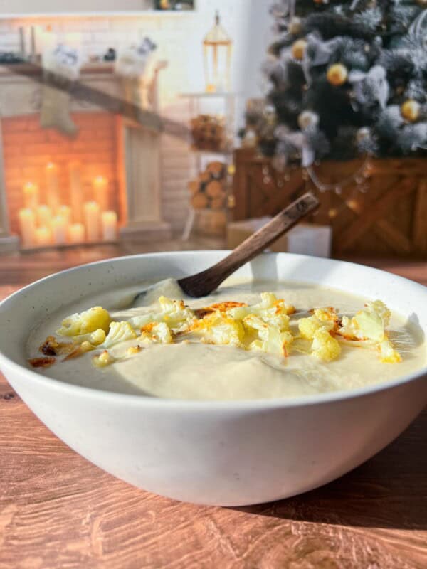 A bowl of creamy soup topped with roasted cauliflower sits on a wooden table. In the background, a cozy room features a decorated Christmas tree and a lit fireplace adorned with candles and seasonal decor.