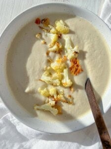 A bowl of creamy cauliflower soup topped with roasted cauliflower florets. A dark wooden spoon rests on the edge of the white bowl. The soup is placed on a white cloth, with sunlight casting a soft shadow.