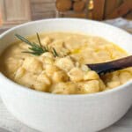 A creamy bowl of gnocchi soup garnished with fresh rosemary. The soup is in a white bowl, and a wooden spoon rests inside. A cozy, rustic background with stacked firewood adds warmth to the scene.