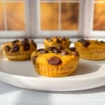 Three pumpkin muffins with chocolate chips on a white plate are placed on a light-colored table. The background shows a window with autumn scenery, featuring warm, golden hues. The muffins are bathed in soft natural light.