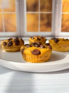 Three pumpkin muffins with chocolate chips on a white plate are placed on a light-colored table. The background shows a window with autumn scenery, featuring warm, golden hues. The muffins are bathed in soft natural light.