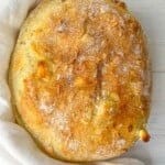 A freshly baked round loaf of bread with a golden, crispy crust rests on a light cloth on a white wooden surface. The bread is dusted with flour, showcasing its rustic texture.