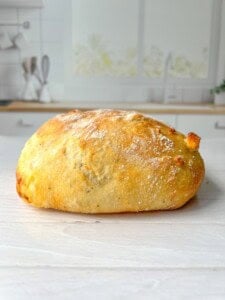 A freshly baked loaf of bread on a light wooden surface in a kitchen setting, with white cabinets and kitchen utensils visible in the blurred background. The bread has a golden, crispy crust and is dusted with flour.