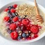 A bowl of oatmeal topped with fresh raspberries, blueberries, a honey drizzle, and a sprinkle of seeds, served with a wooden honey dipper on a light wooden table.