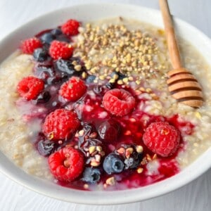 A bowl of oatmeal topped with fresh raspberries, blueberries, a honey drizzle, and a sprinkle of seeds, served with a wooden honey dipper on a light wooden table.